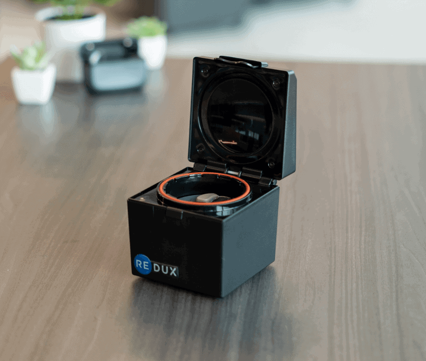 A small black RE DUX electronic device with an open lid is placed on a wooden table. The background is slightly blurred, showing small potted plants and another electronic device.