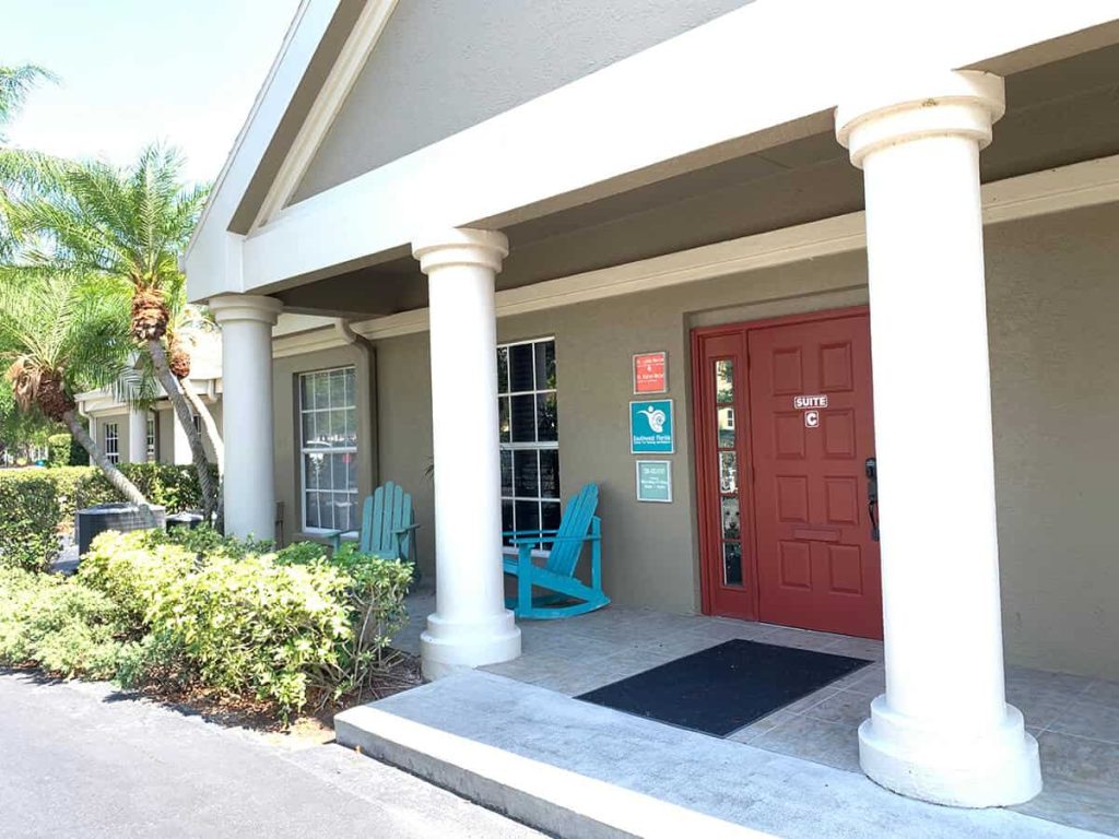 A building entrance with white columns, a red door marked SUITE C, signboards, two turquoise Adirondack chairs, windows, and surrounding greenery, including bushes and a palm tree.