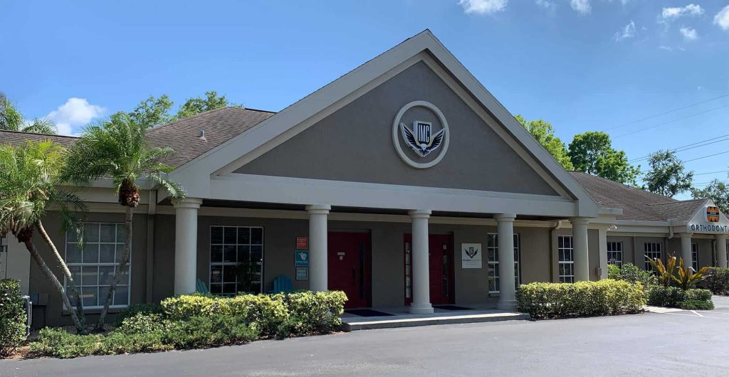 A single-story building with a triangular roof, columns, and red double doors. Shrubs and palm trees line the entrance, and a circular emblem is displayed above the doors. The sky is blue with some clouds.