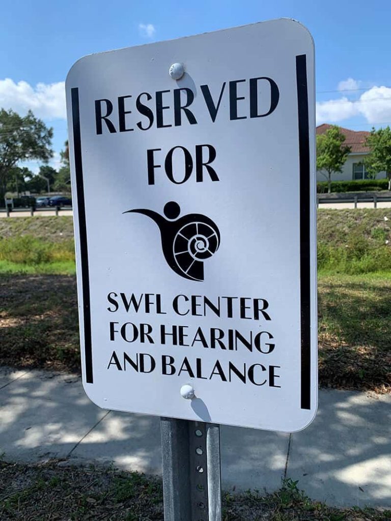 A white sign reads Reserved for SWFL Center for Hearing and Balance with a stylized ear and spiral logo, situated outdoors near a sidewalk and grassy area.