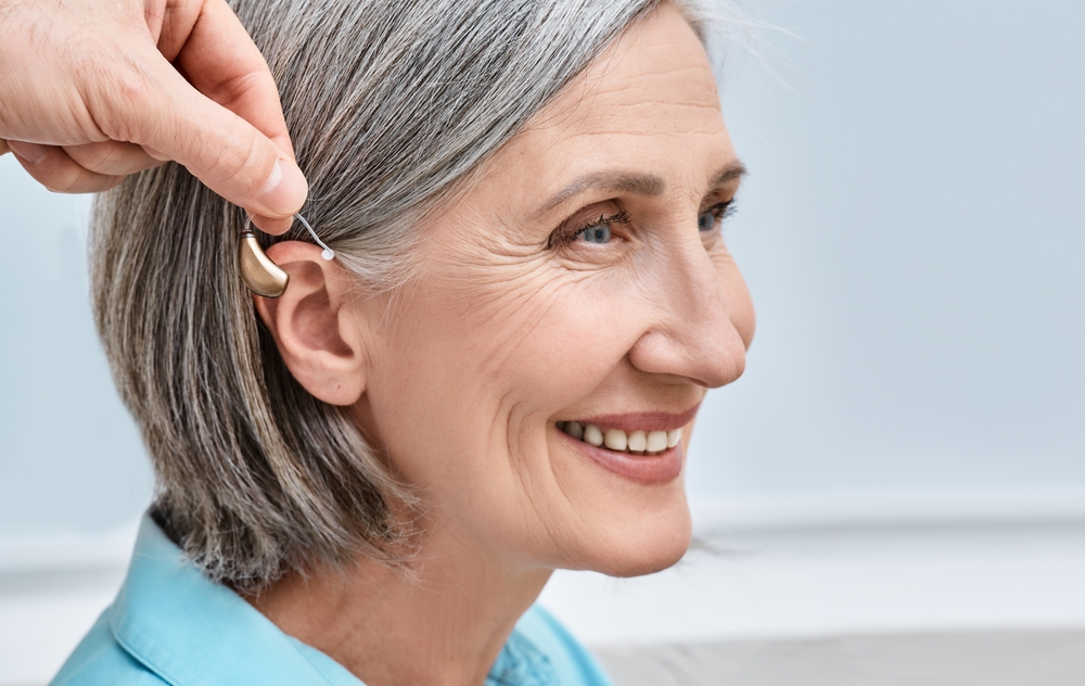 A smiling older woman with gray hair is having a hearing aid placed behind her ear by another person.
