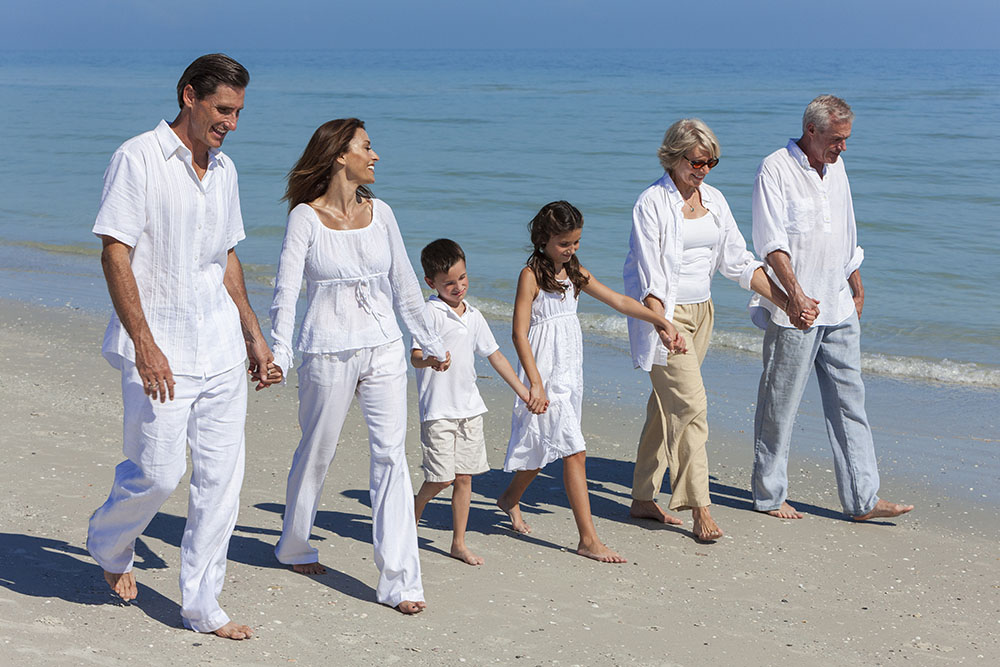 Three generations of a family, dressed in white, walk barefoot along a sandy beach by the ocean on a sunny day, smiling and holding hands.