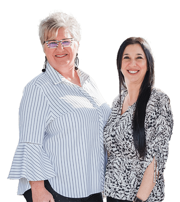 Two women standing side by side, smiling at the camera. One has short gray hair and glasses, wearing a striped shirt; the other has long dark hair and is wearing a patterned blouse. The background is plain white.