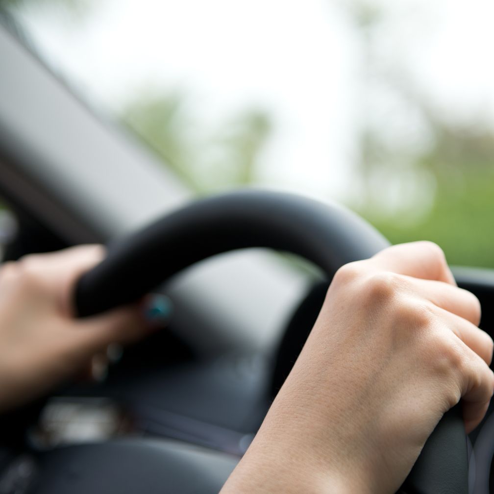 Close-up of a person’s hands gripping a black steering wheel while driving a car. The background is blurred, showing greenery outside the vehicle window.