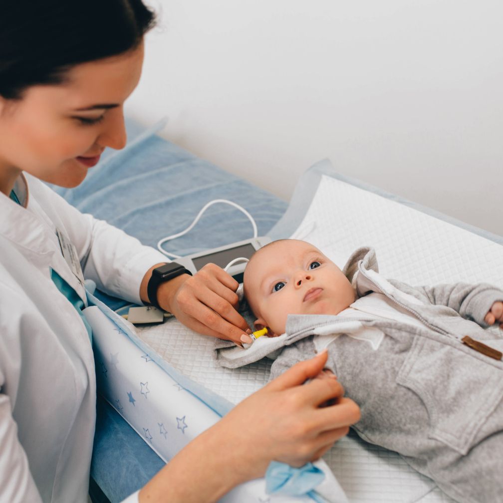 A healthcare professional tests a babys hearing by placing small earphones in the infants ears as the baby lies on a changing table. The baby looks up calmly while the clinician gently conducts the examination.