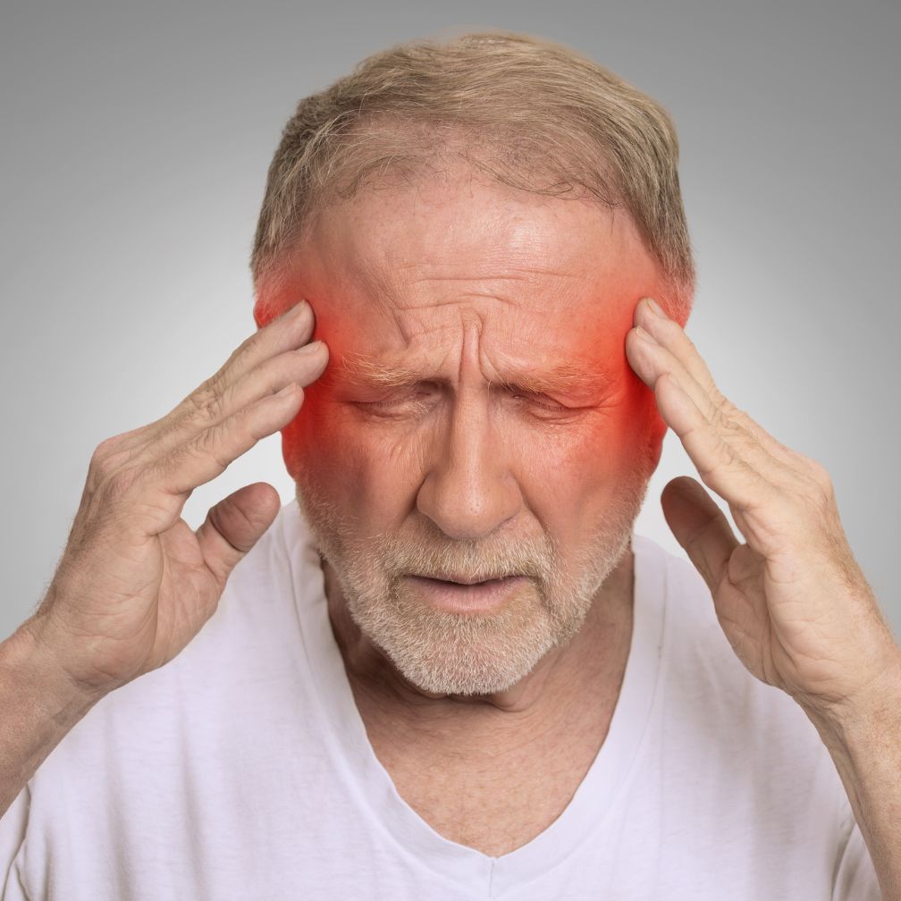 Older man with closed eyes presses his temples, looking pained. Red shading highlights the sides of his head, indicating a headache or migraine. He wears a white shirt and stands against a gray background.